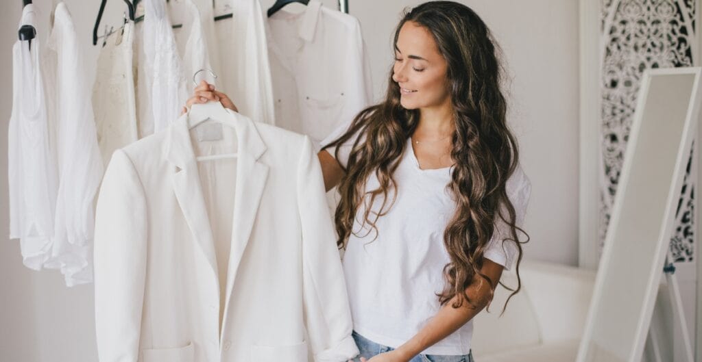A woman with long, curly hair is smiling as she holds up a white blazer on a hanger in front of a mirror. She is standing in a clothing store or boutique with white garments hanging on a rack behind her.