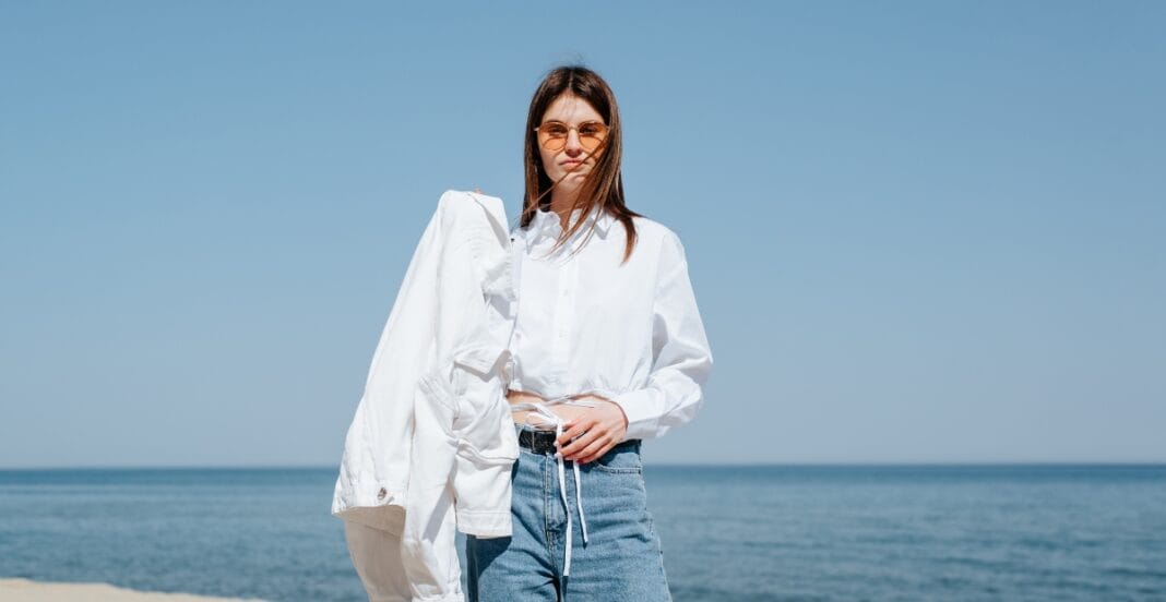A young woman standing on a beach with the ocean and clear blue sky in the background. She is wearing sunglasses, a white shirt, and blue jeans, holding a white jacket over her shoulder.