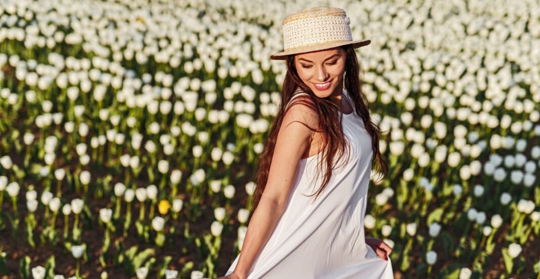 A woman with long brown hair, wearing a white dress and a straw hat, standing in a field of blooming white flowers, smiling and looking down.
