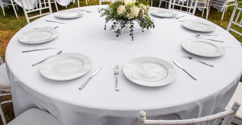 A round banquet table set for a formal outdoor event with white plates, silver cutlery, and a floral centerpiece, surrounded by white chairs on a grassy area.