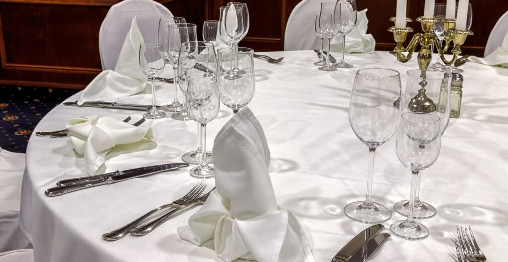 Elegant round dining table set with a white tablecloth, multiple wine glasses, neatly folded napkins, and polished silverware, with a gold candelabrum and white chairs in the background.
