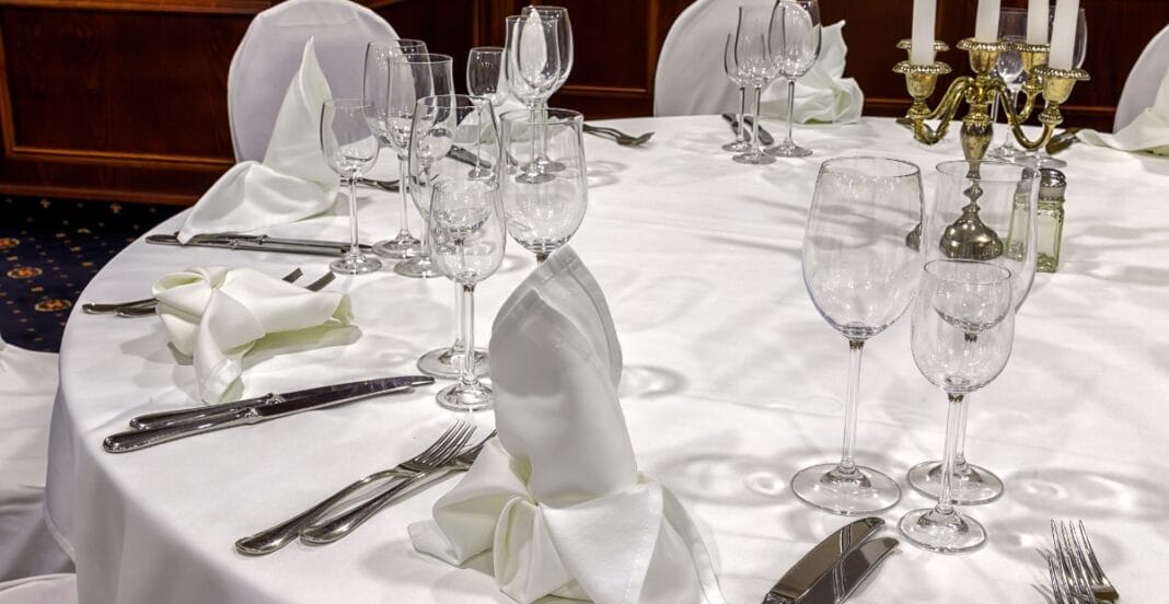 Elegant round dining table set with a white tablecloth, multiple wine glasses, neatly folded napkins, and polished silverware, with a gold candelabrum and white chairs in the background.