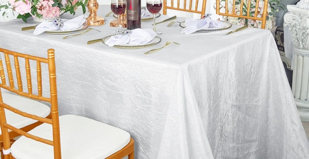 A close-up of a formally set dining table with a white textured tablecloth, gold flatware, white napkins, wine glasses, and a floral centerpiece. Golden chairs with white cushions surround the table.