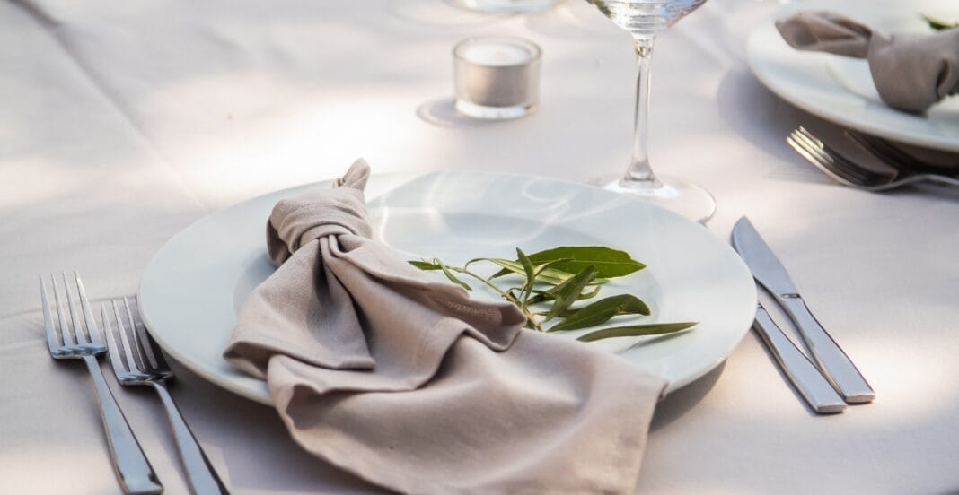A elegantly set dining table with a white plate, beige cloth napkin decorated with green leaves, silver cutlery, a wine glass, and a small candle in the background.