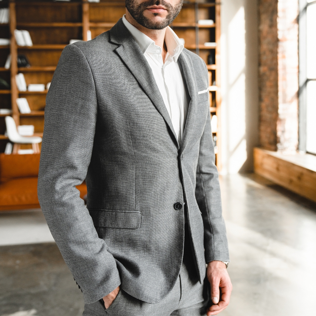  A well-dressed man wearing a grey sport coat and white shirt standing indoors, with a background featuring wooden shelves and a large window.