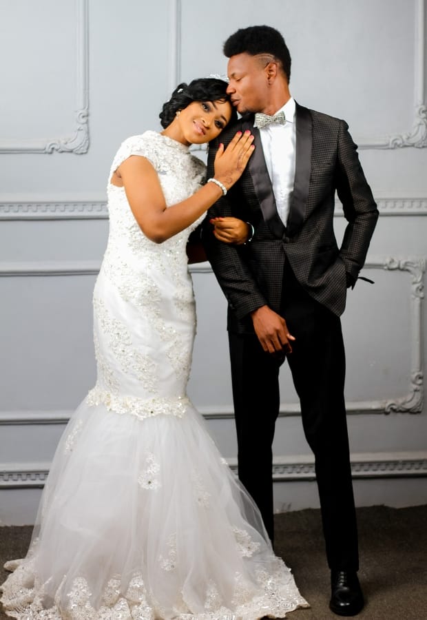 A bride and groom pose closely together, with the bride wearing a white mermaid wedding gown featuring lace detailing and a fitted bodice that flares out at the hem, and the groom in a black tuxedo with a textured pattern and a bowtie.