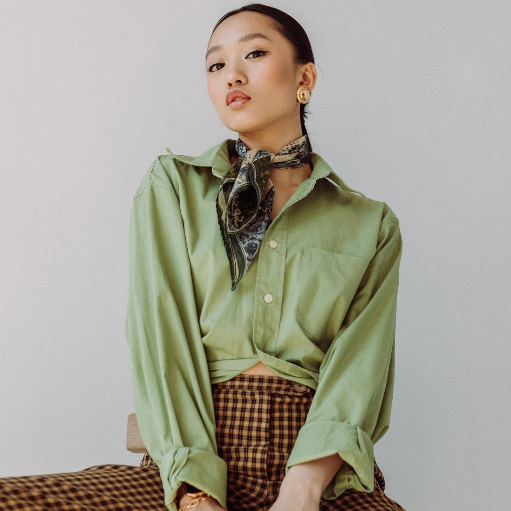 A stylish young woman with dark hair pulled back, wearing a light green oversized button-up shirt, a patterned scarf tied around her neck, gold earrings, and checkered brown and black pants, sitting against a plain light background with a confident expression.