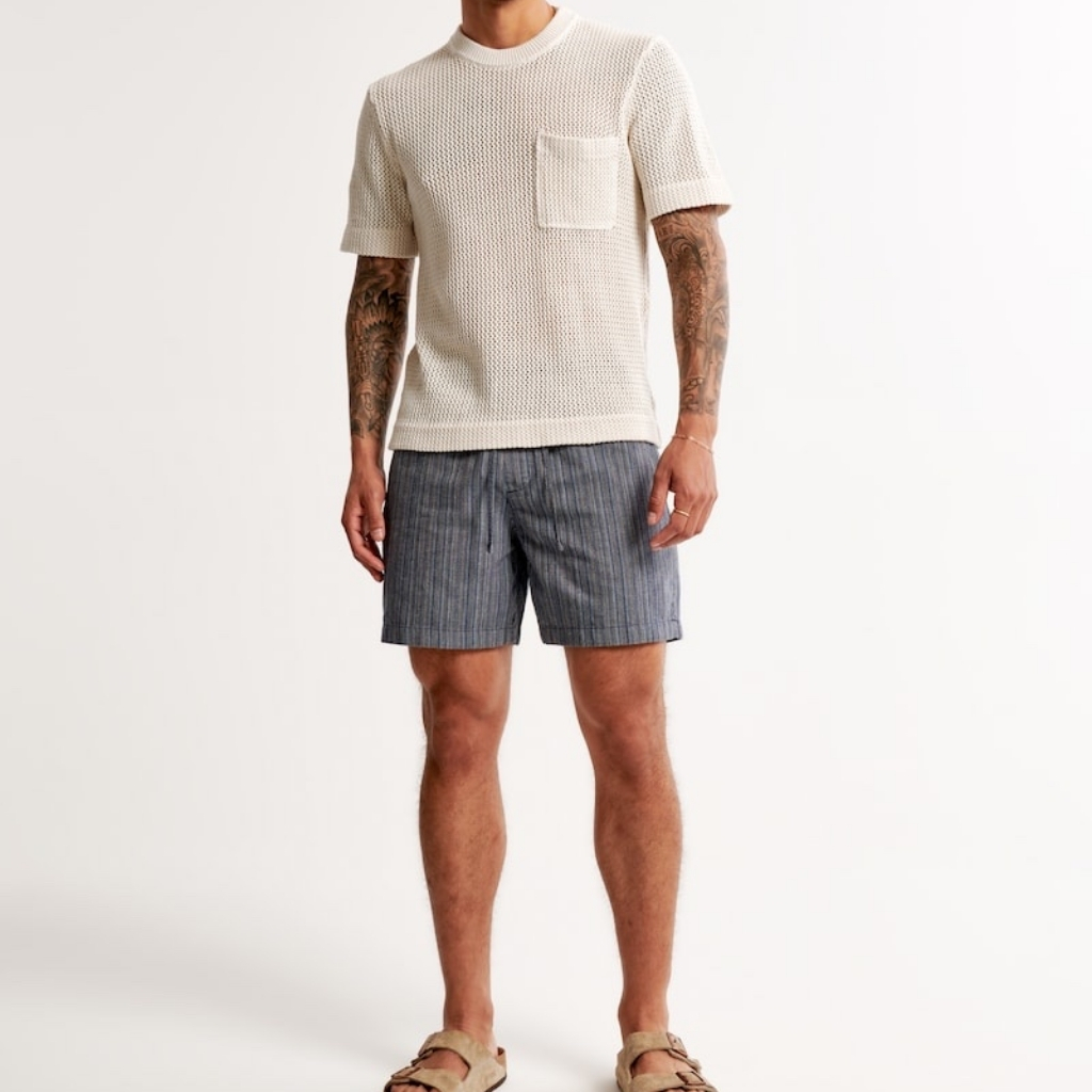 A person wearing a cream-colored knitted short-sleeve top with a pocket on the chest, paired with loose-fitting gray striped shorts and beige sandals, standing against a plain white background.