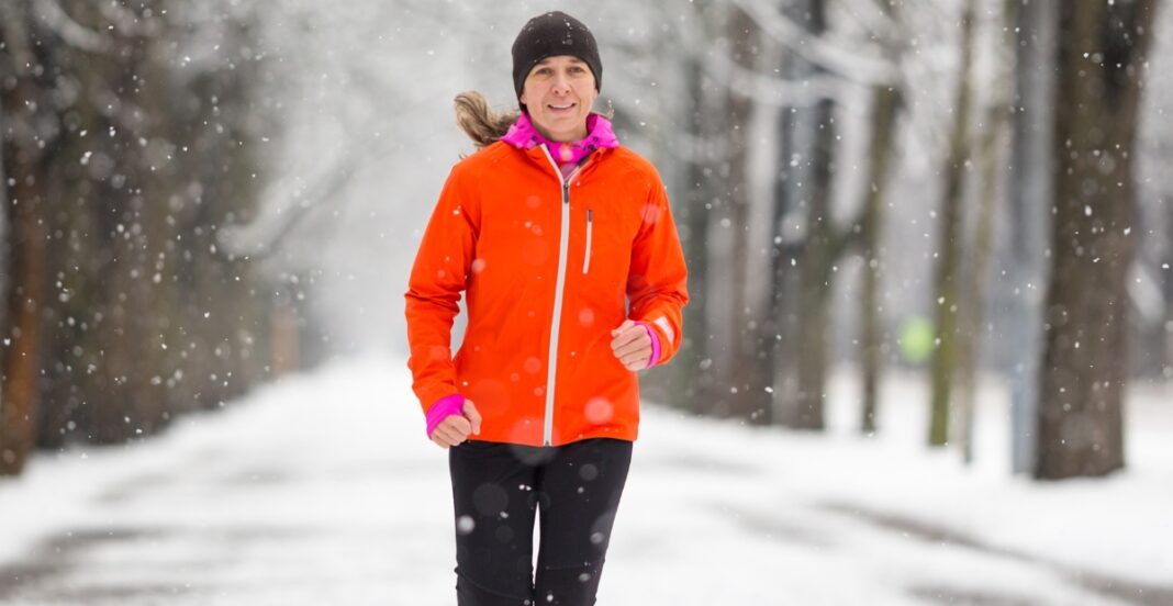 A woman jogging outdoors in winter weather, wearing a bright orange winter jacket with pink accents, black running pants, a black beanie, and gloves, with snow falling around her.