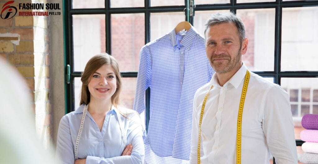 A fashion designer and a client are standing in a bright studio with large windows, smiling at the camera. Behind them, a blue checkered shirt is hanging on a hanger, and fabric rolls are visible on the right side.