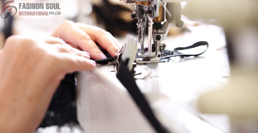 Close-up of hands guiding black fabric through a sewing machine, with the sewing machine in focus and the person's hands slightly blurred. The image shows a sewing process, likely in a textile or fashion workshop, with the logo 
