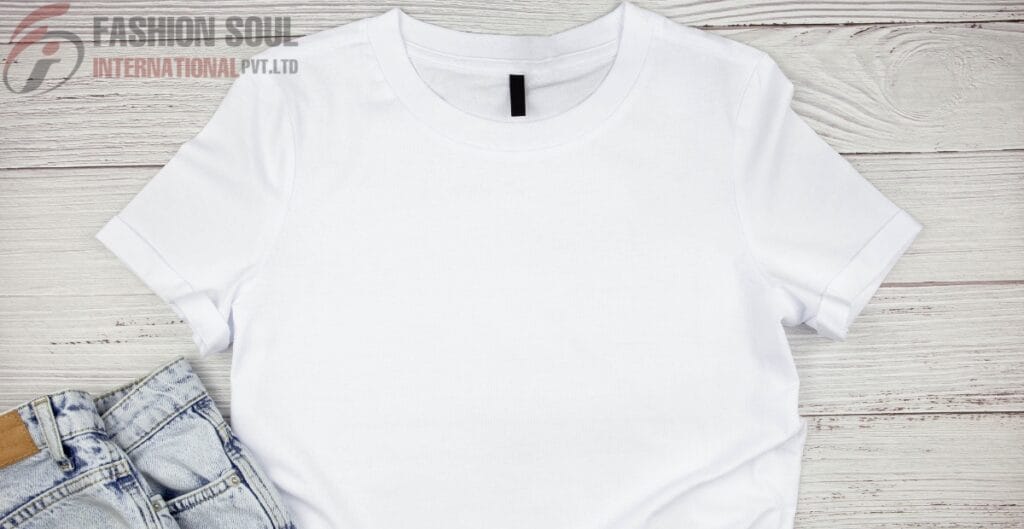 A plain white T-shirt laid flat on a light-colored wooden surface, partially covered by a pair of light-wash denim jeans in the bottom left corner.