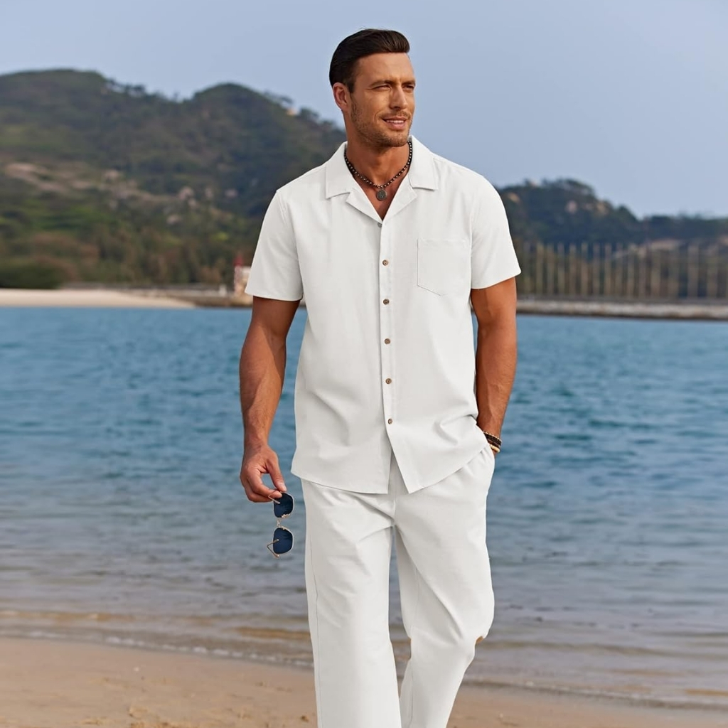  A man dressed in a white short-sleeved shirt and matching white trousers stands barefoot on a beach, holding a pair of sunglasses in his right hand. He is looking slightly to the side with a relaxed expression, with a scenic background of water, green hills, and a cloudy sky.