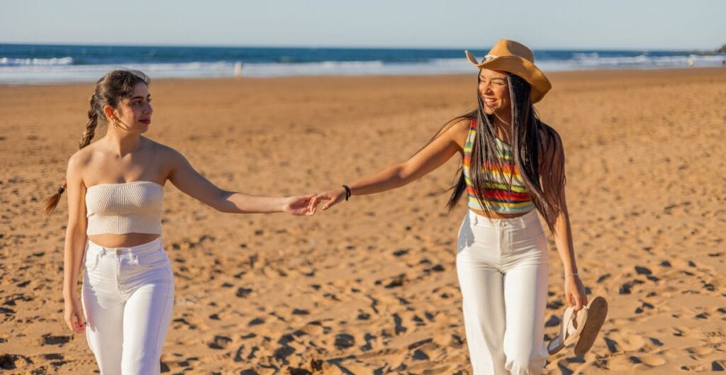 Two women walking hand-in-hand on the beach, smiling, with the ocean and clear sky in the background.
