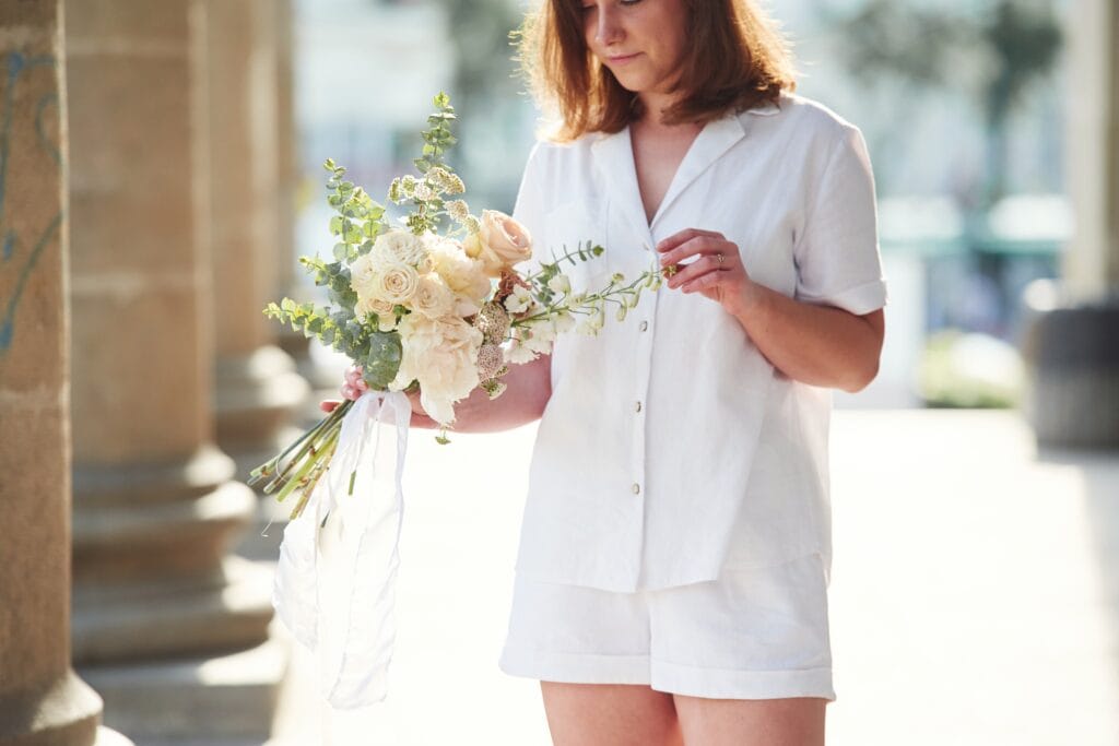 A woman dressed in white, holding a bouquet of cream and pink flowers, standing outdoors near stone columns.