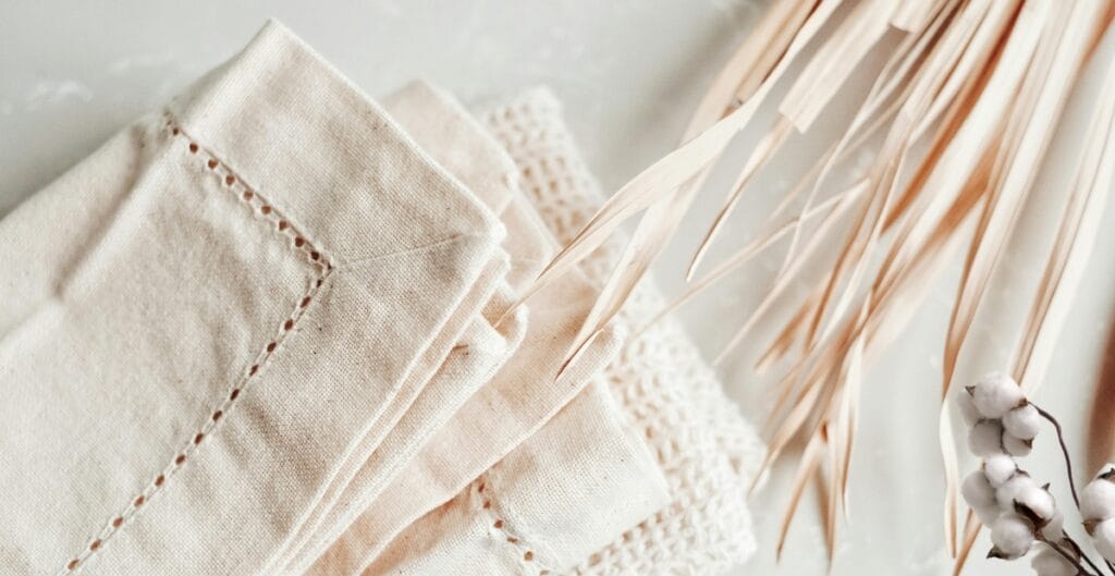 A close-up photograph of neatly folded beige fabric, likely linen or cotton napkins, placed on a surface with a sprig of dried pampas grass and some cotton bolls nearby. The scene has a soft, neutral, and minimalist aesthetic.