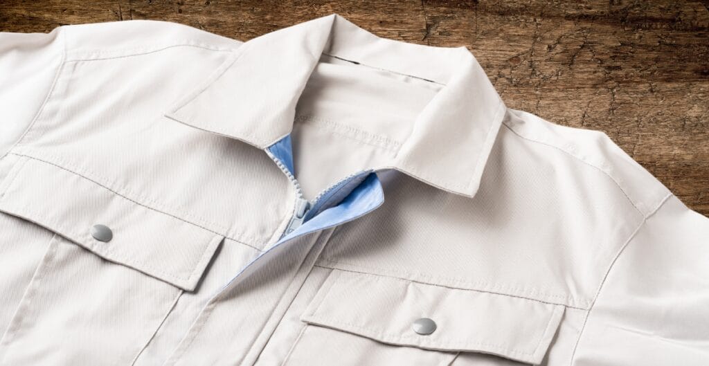 A close-up of a white Western-style jacket with snap buttons, lying on a wooden surface. The jacket has epaulets on the shoulders and a concealed zipper underneath the front flap.