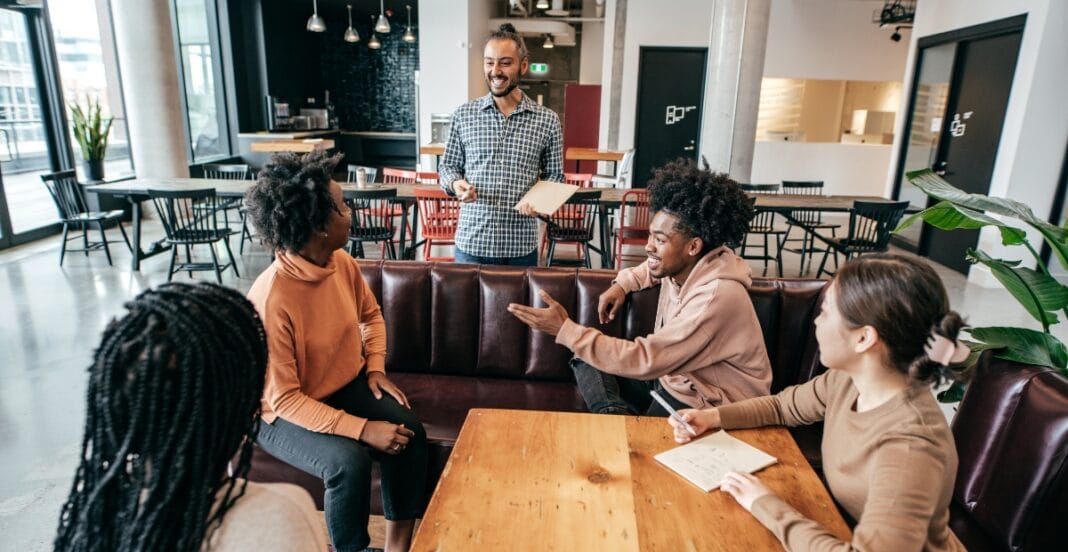 A group of four diverse young adults engaged in a lively discussion in a modern, well-lit café or coworking space. One person is standing and holding a notebook, while the other three are seated around a wooden table, actively interacting and smiling.