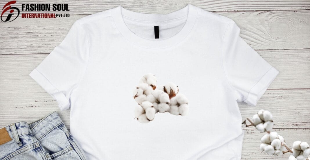White T-shirt with cotton plant print in the center, laid flat on a light wooden surface next to denim shorts and a cotton branch.