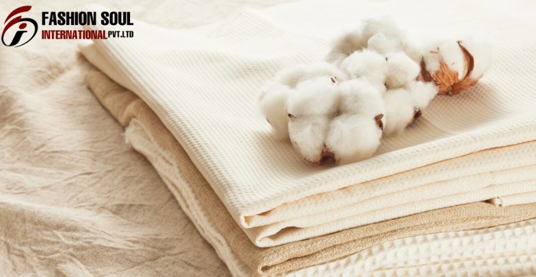 A neatly folded stack of neutral-toned fabrics with a piece of cotton and a cotton boll placed on top, against a textured background. The top left corner features the logo and text for 