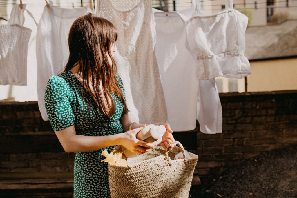 A woman with long brown hair, wearing a green dress with white floral patterns, is holding a bottle while standing outdoors near clothes hanging to dry on a laundry line.