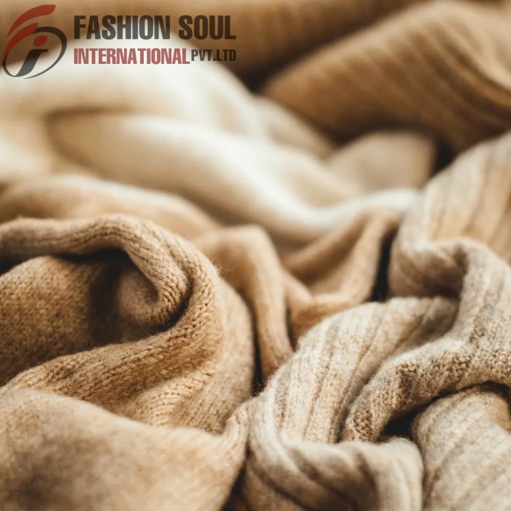 Close-up photograph of soft, beige, and brown fabric or clothing materials arranged in folds, with the logo and text "FASHION SOUL INTERNATIONAL PVT. LTD" in the top left corner.