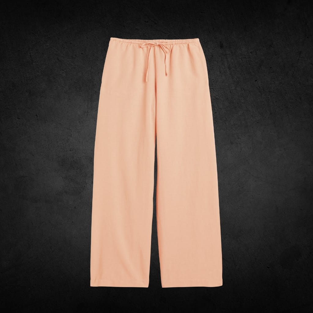 A pair of light peach-colored wide-leg pants with an elastic waistband and drawstring, displayed against a dark textured background.
