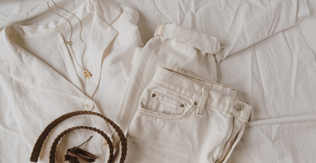 A neatly arranged flat lay of cream-colored clothing and accessories on a soft surface, including a button-up shirt with a necklace, a pair of pants, a belt, and eyewear.