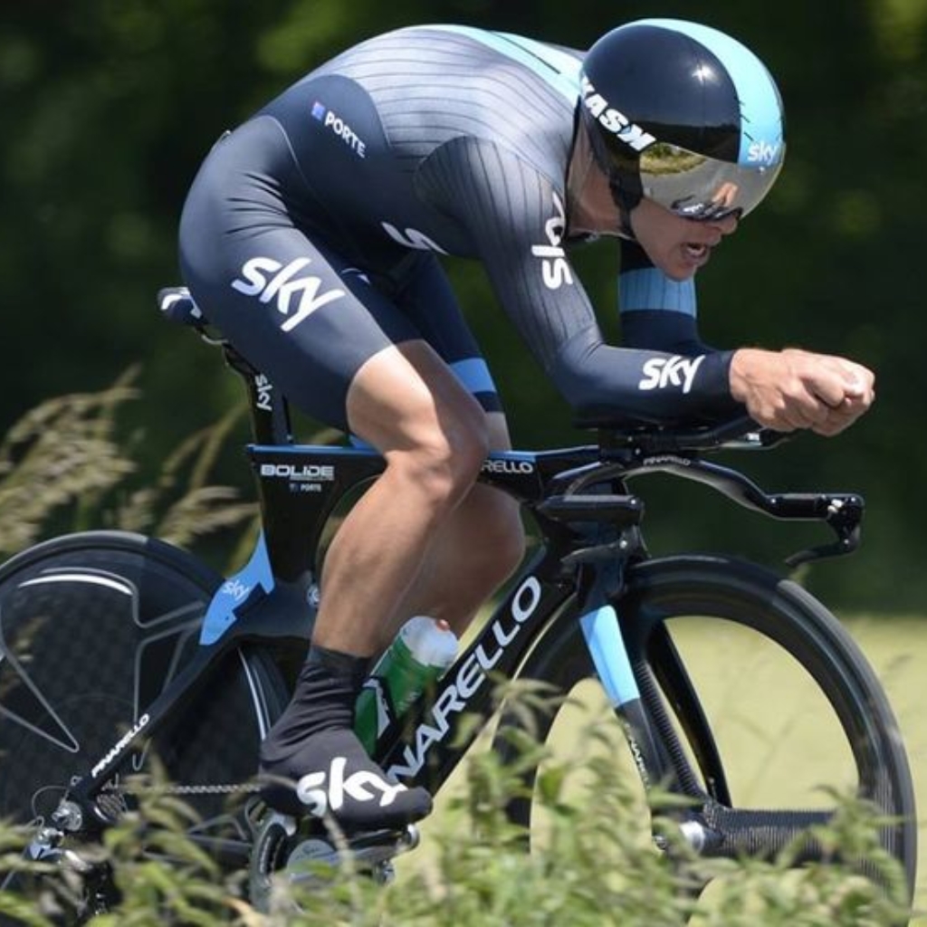 A competitive cyclist wearing a SKY Sports team uniform, helmet, and sunglasses, riding a black and blue bicycle outdoors on a grassy area.