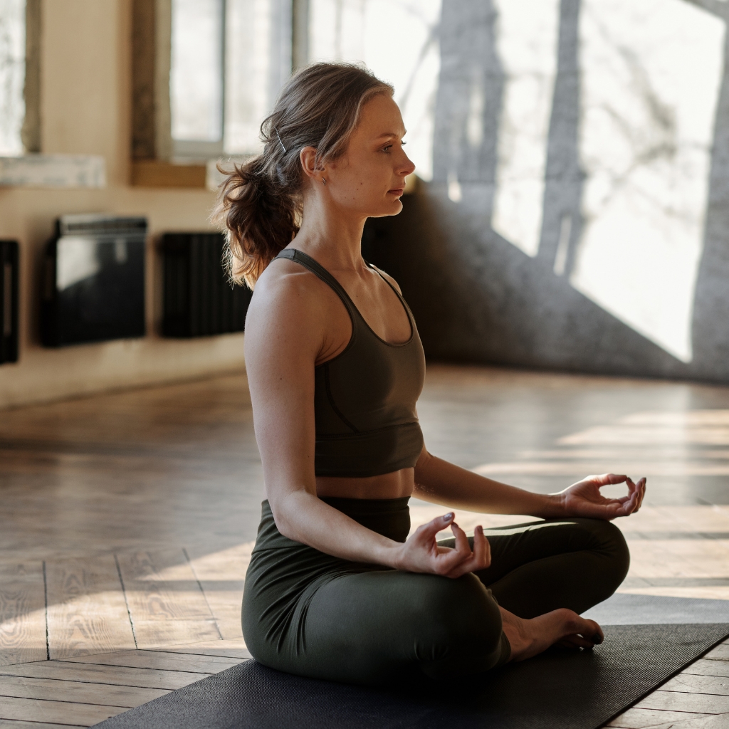 A woman practicing yoga indoors, sitting cross-legged on a yoga mat, wearing a dark sports bra and matching leggings.
