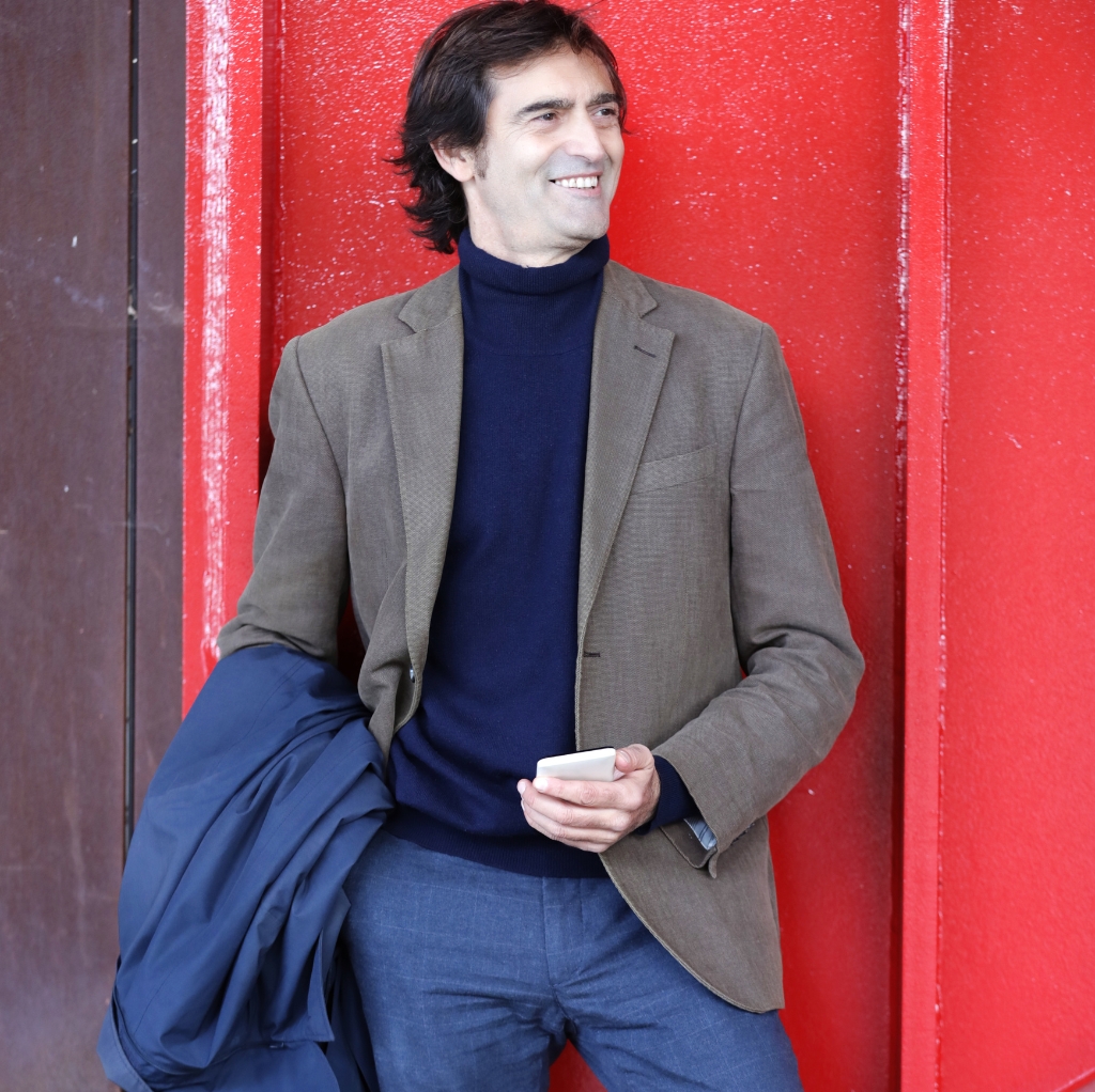 A man wearing a navy blue turtleneck sweater paired with a brown sport coat, holding a jacket in his right hand, standing against a red wall and smiling.