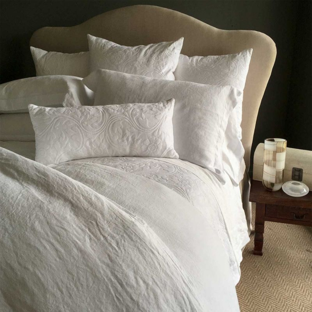 A luxurious bed with an ornate beige headboard, dressed in white embroidered bedding and multiple white pillows, alongside a wooden nightstand with a ceramic vase and decorative object.