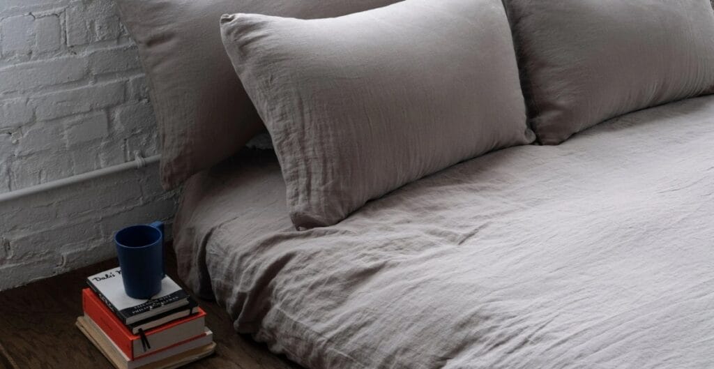 A neatly made bed with light gray bedding and pillows, positioned next to a small side table holding a stack of books and a blue cup, against a white brick wall.