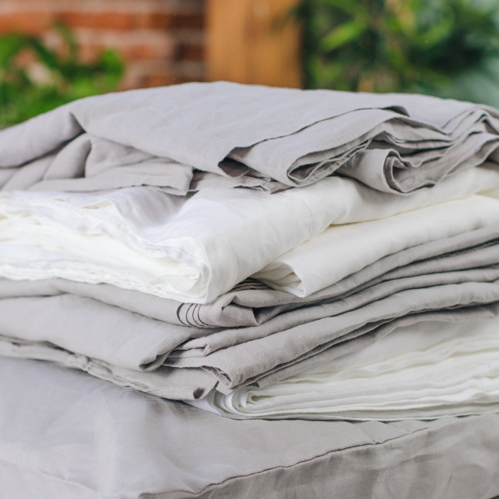 A neatly folded stack of white and light gray linen and fabric, set outdoors with a blurred background featuring greenery and brickwork.
