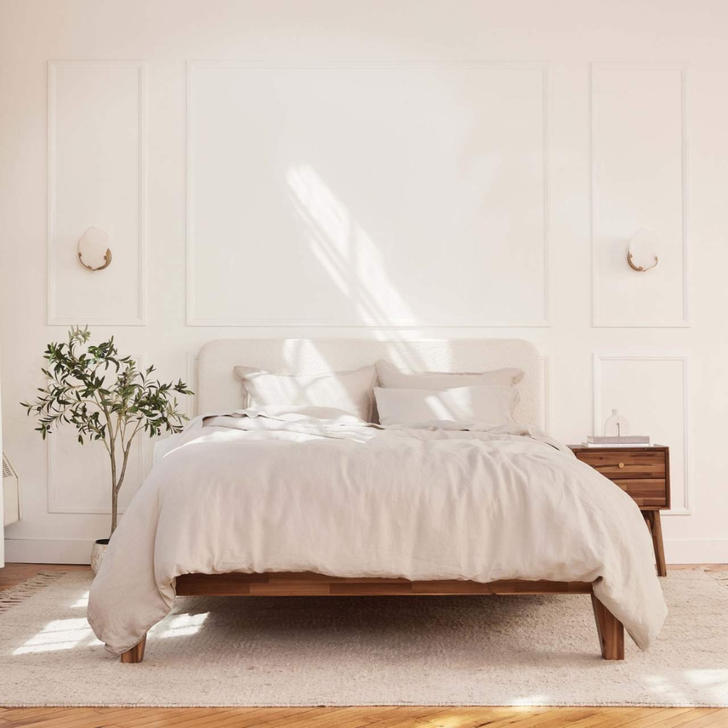 A minimalist bedroom with a large bed dressed in white linens, a wooden nightstand on the right, a potted plant on the left, and soft natural light streaming through a window, casting shadows on the wall.