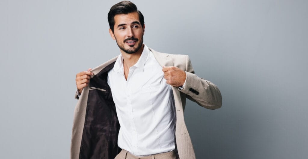 A confident man with dark hair and a trimmed beard, wearing a light-colored suit jacket over a white shirt, is looking at the camera and showing off the inside of his sport coat.