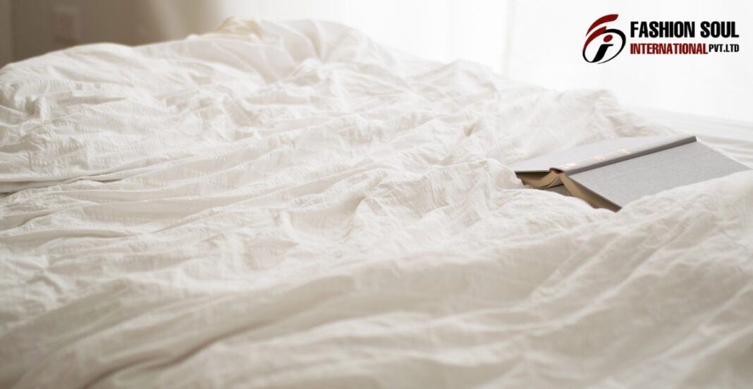 A close-up of a white bed with crumpled bedsheets and an open book resting on it. There is a logo in the top right corner that reads 