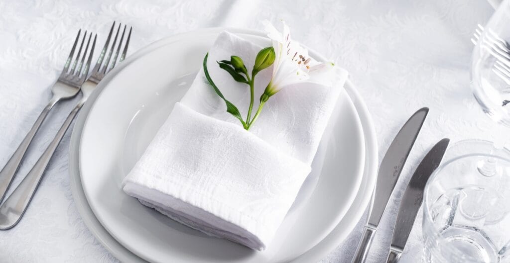 A neatly arranged table setting with a white plate holding a napkin folded with a small white flower and green leaves, surrounded by silverware and clear glasses on a white tablecloth.