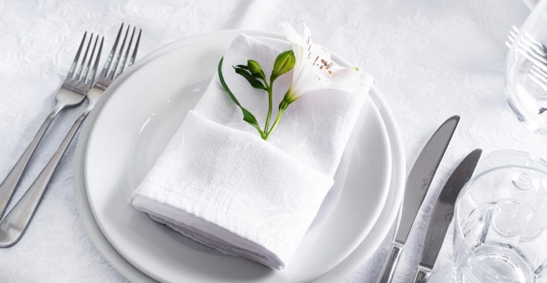 A neatly arranged table setting with a white plate holding a napkin folded with a small white flower and green leaves, surrounded by silverware and clear glasses on a white tablecloth.