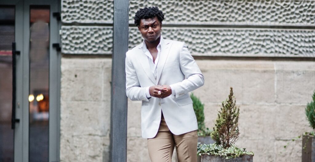 A man with curly black hair wearing a white blazer and beige pants stands outdoors, leaning against a pole with a serious expression.