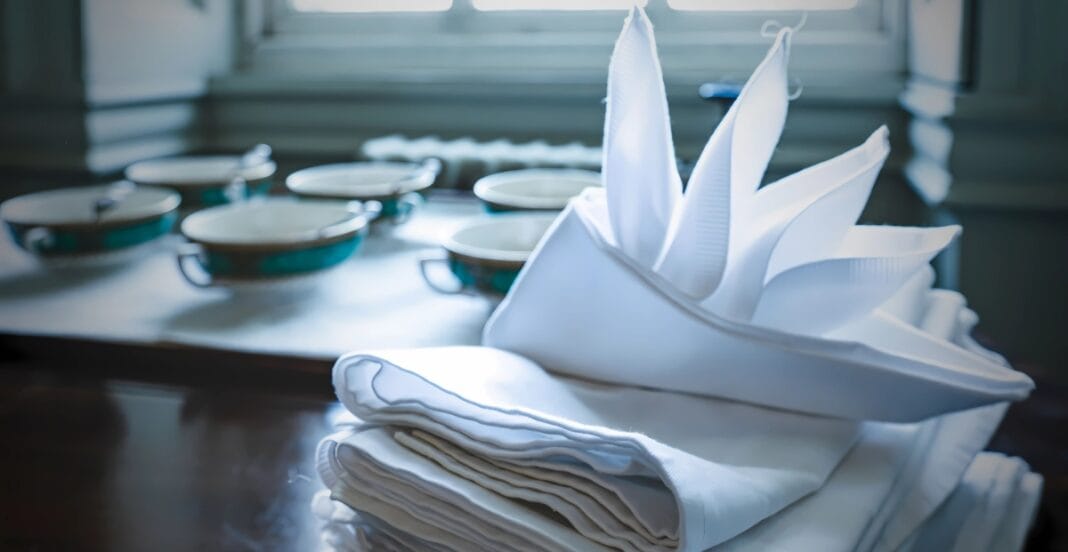 A neatly folded set of white napkins with a decorative display, placed in front of a tray of teacups and saucers on a wooden table near a window.