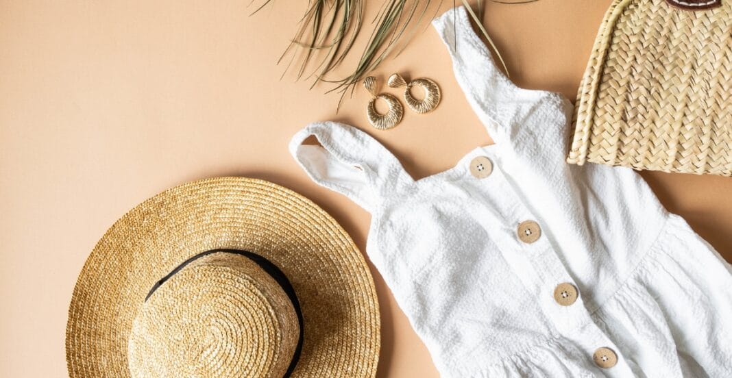 A flat lay of summer accessories on a light background, including a wide-brimmed straw hat, a white sleeveless dress with wooden buttons, gold hoop earrings, and a woven straw bag.