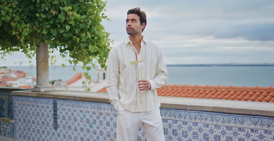 A man with dark hair and a beard stands outdoors near a tiled railing, holding a flower in his right hand and looking thoughtfully to his left. The background features a tree, rooftops, and a body of water under a cloudy sky.