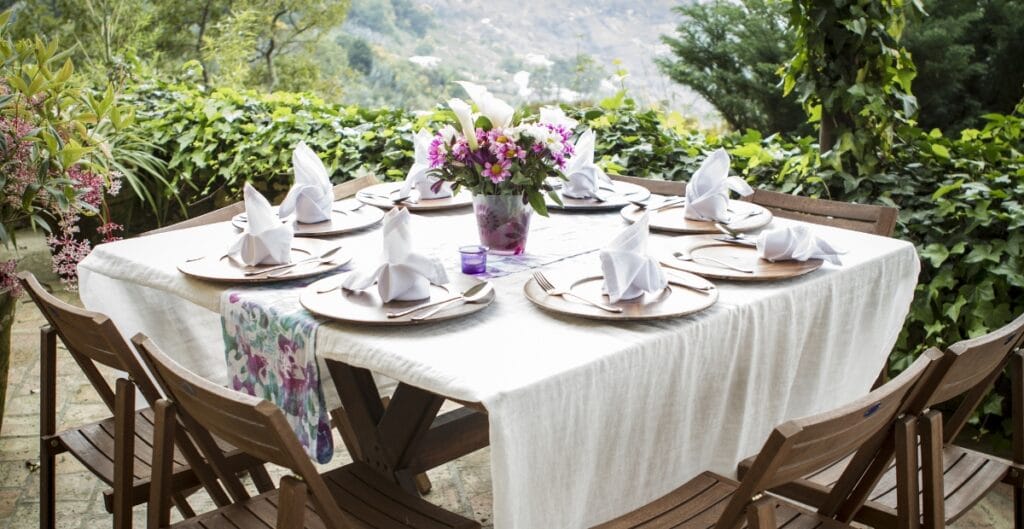 An outdoor dining table set with nine white place settings, each with a neatly folded white napkin, surrounded by wooden chairs, with a lush green garden in the background.