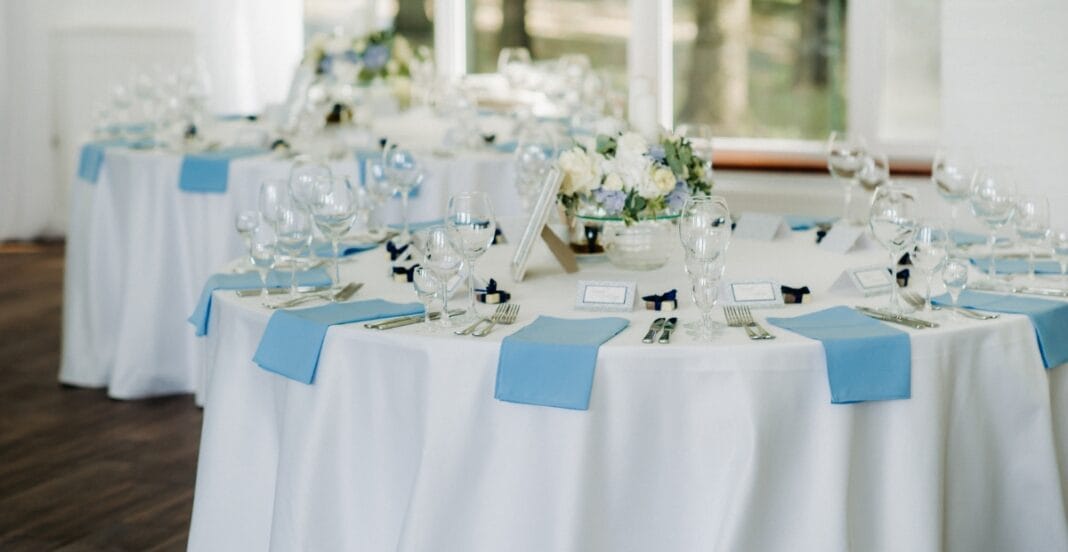 Elegant round banquet table set for a formal event, decorated with white tablecloths, light blue napkins, multiple wine glasses, and a floral centerpiece, with natural light coming through the windows.