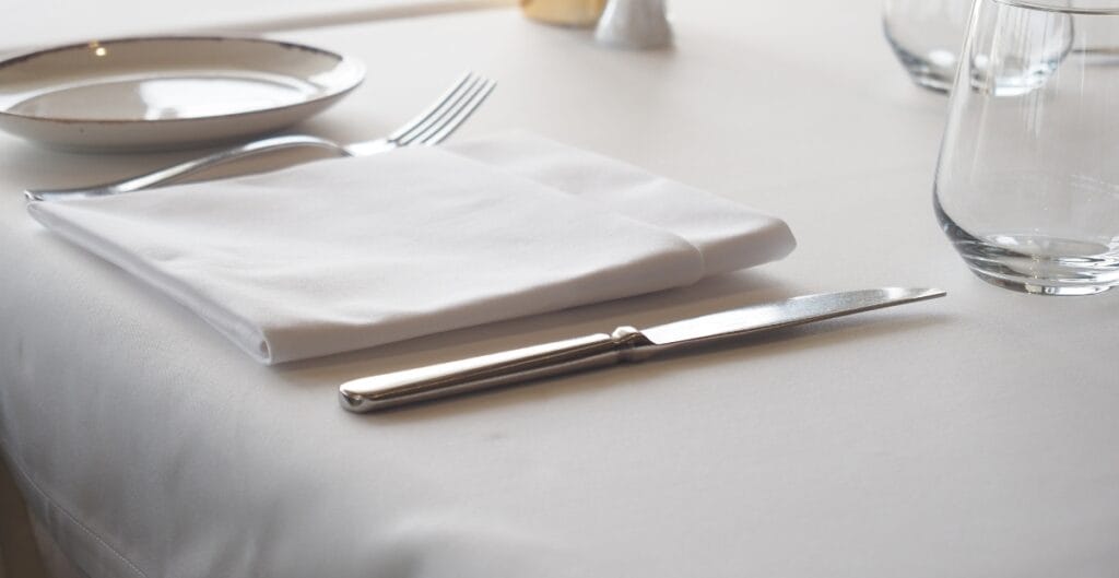 A neatly set dining table with a white tablecloth, a folded white napkin, a knife, a fork, a plate, and a glass of water.