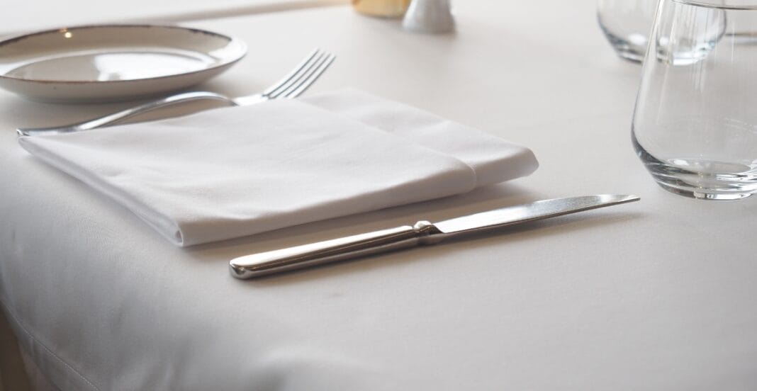 A neatly set dining table with a white tablecloth, a folded white napkin, a knife, a fork, a plate, and a glass of water.