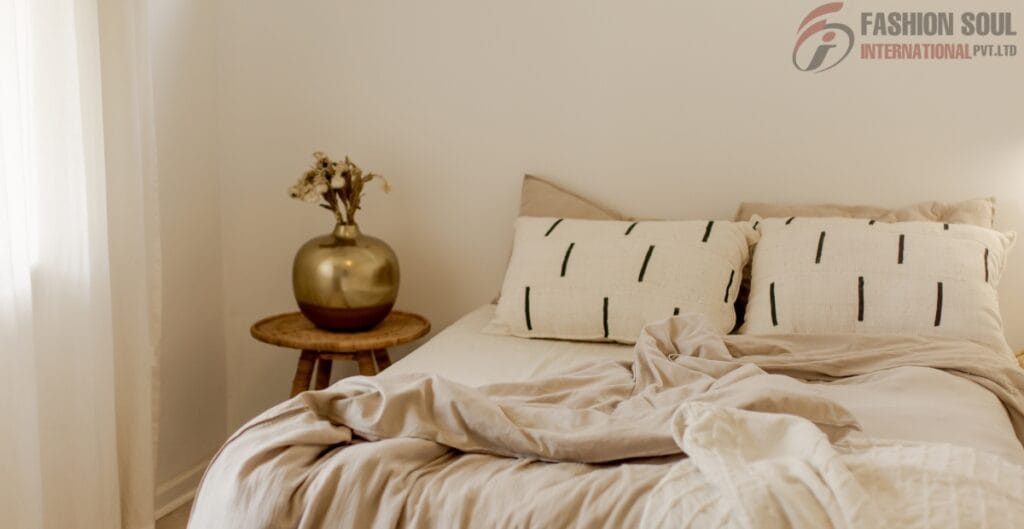 A neatly made bed with beige and cream bedding, two striped pillows, and an abstract bed cover, next to a small wooden nightstand holding a metallic gold vase with dried flowers, in a softly lit bedroom.