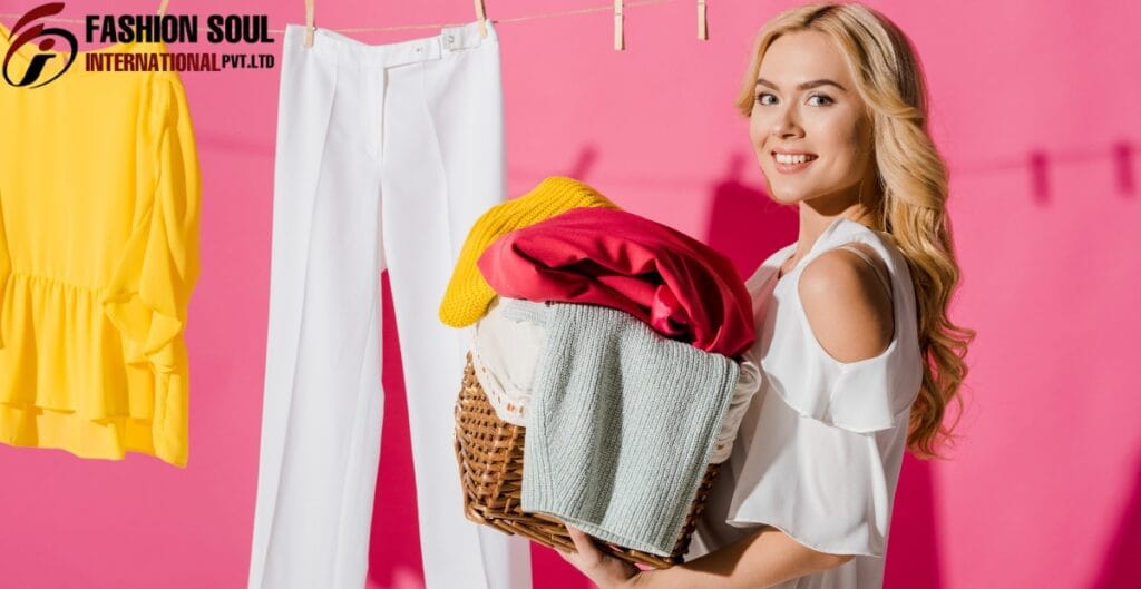 A young woman with blonde hair holding a laundry basket filled with colorful clothes, smiling at the camera. In the background, there are hanging white pants, a yellow top, and a pink wall with clothespins.
