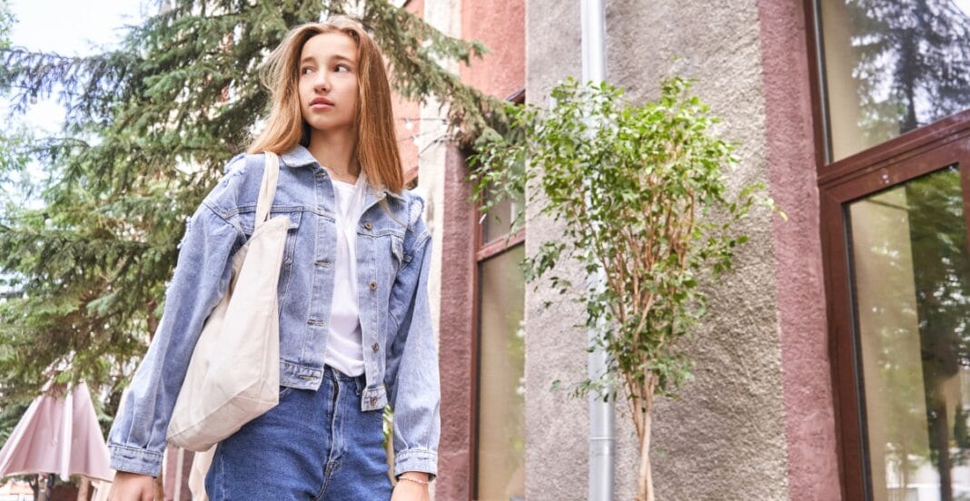A young girl with long hair wearing a denim jacket and jeans, standing outdoors near a building with large windows and some greenery.